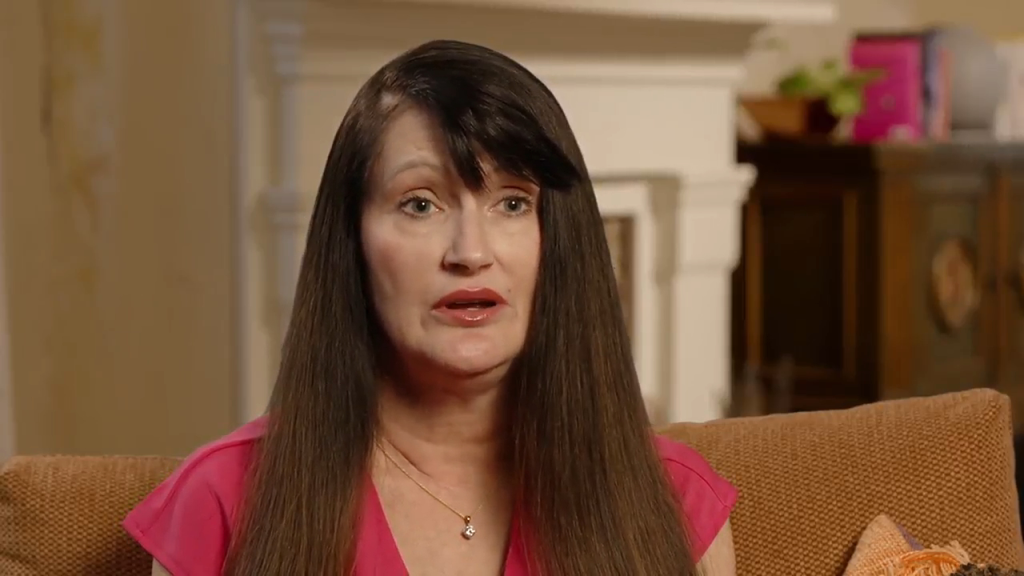 A woman with long dark hair, wearing a pink top and a necklace, speaks while sitting on a brown patterned couch.