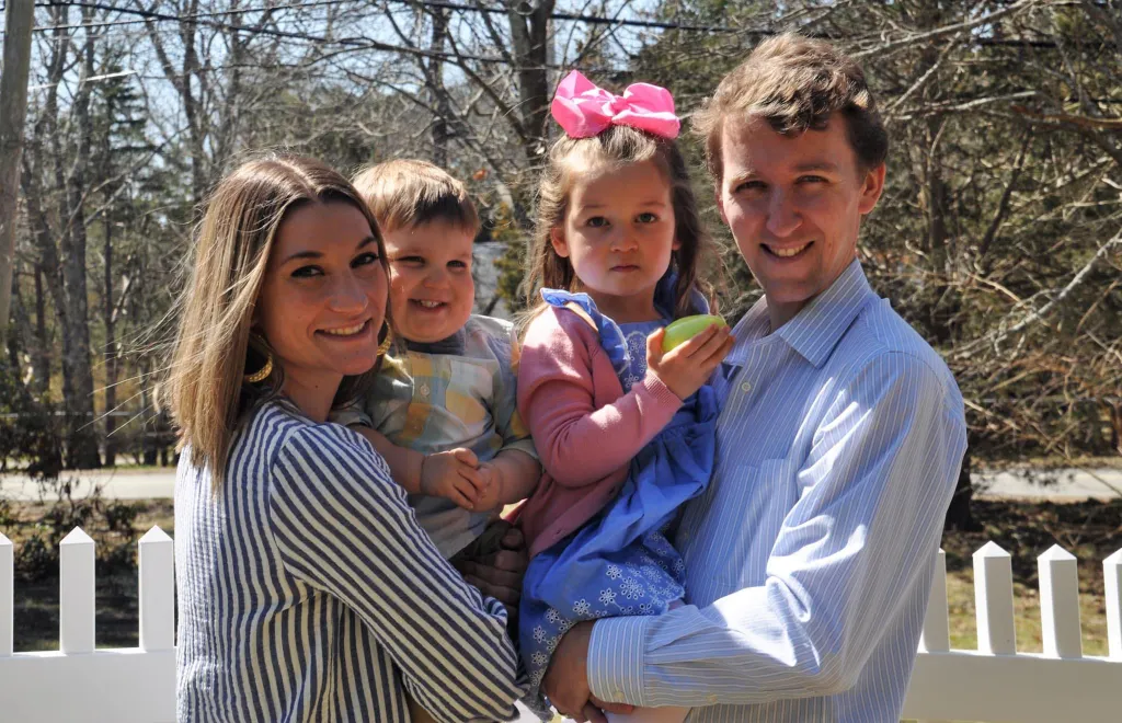 A smiling family of four: a woman holding a baby, and a man holding a little girl with a pink bow in her hair and an Easter egg in her hand.