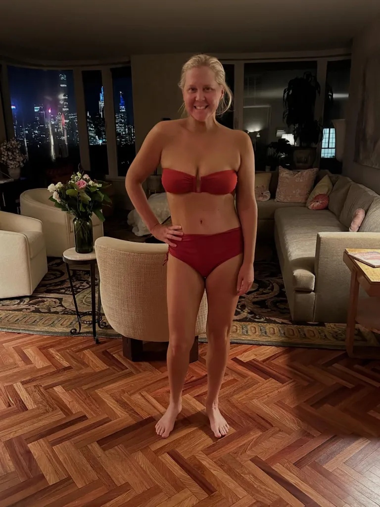 Amy Schumer smiling in a red bikini in her living room, with a city skyline visible through large windows at night.