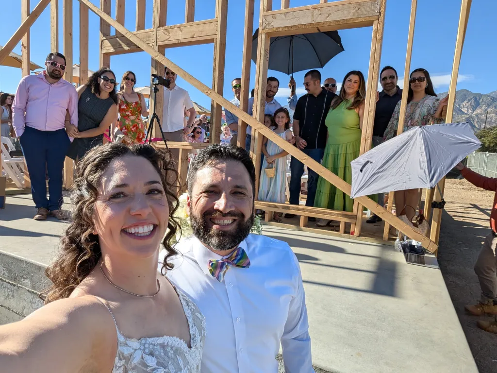 Morgan Soloway and Kenny Rotter smile at the camera as wedding guests stand behind them within the wooden frame of a house.