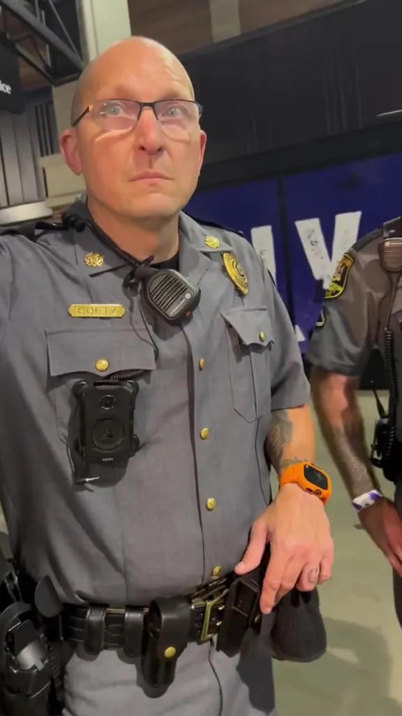 A male police officer with the name "GOETZ" on his uniform, wearing glasses and a body camera, looking upwards.