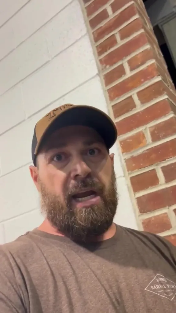 A man with a beard, wearing a baseball cap and a brown t-shirt, looking directly at the camera with a surprised or distressed expression.