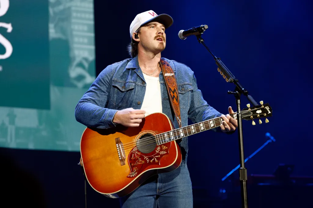 Morgan Wallen performing live on stage, wearing a denim jacket and white baseball cap, holding an acoustic guitar.
