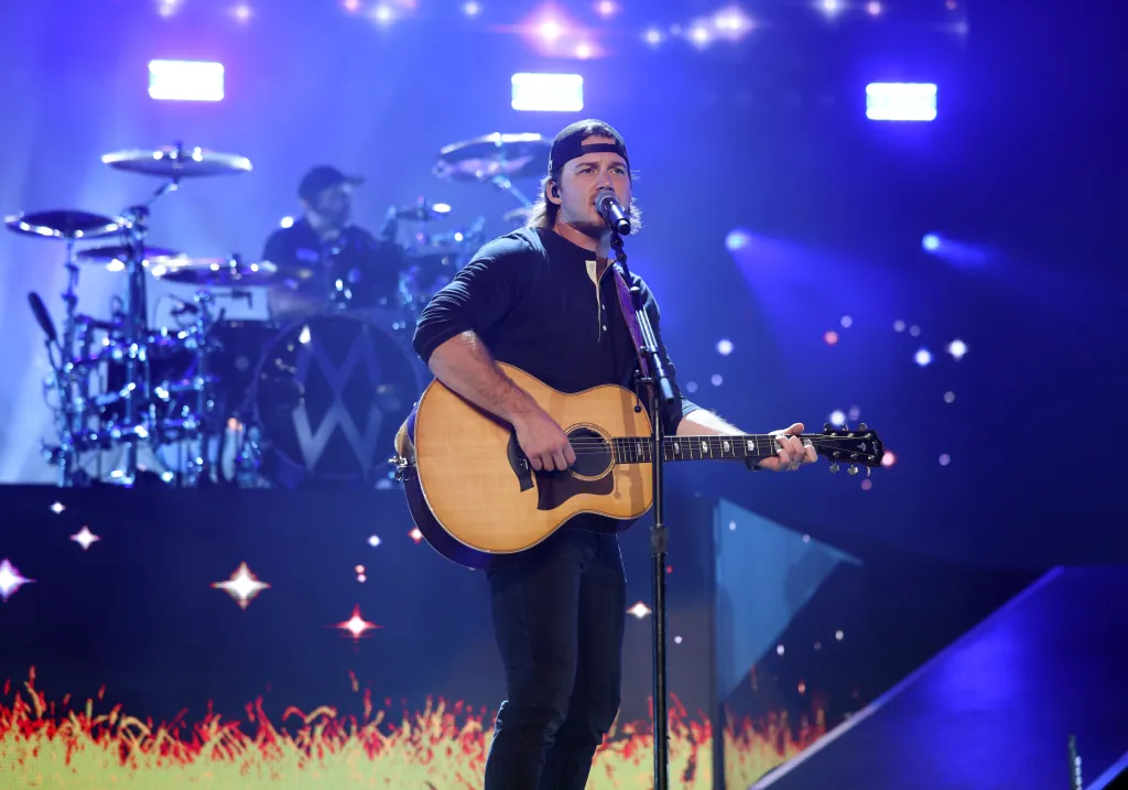 Morgan Wallen singing and playing an acoustic guitar on stage with a drummer in the background.