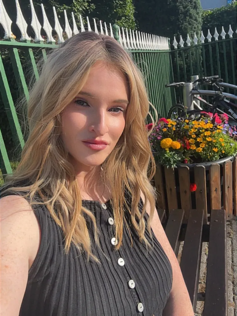 Jamey Carney, a woman with long blonde hair, wearing a black ribbed top, sits on a wooden bench next to a flower planter, with a green fence and trees in the background.