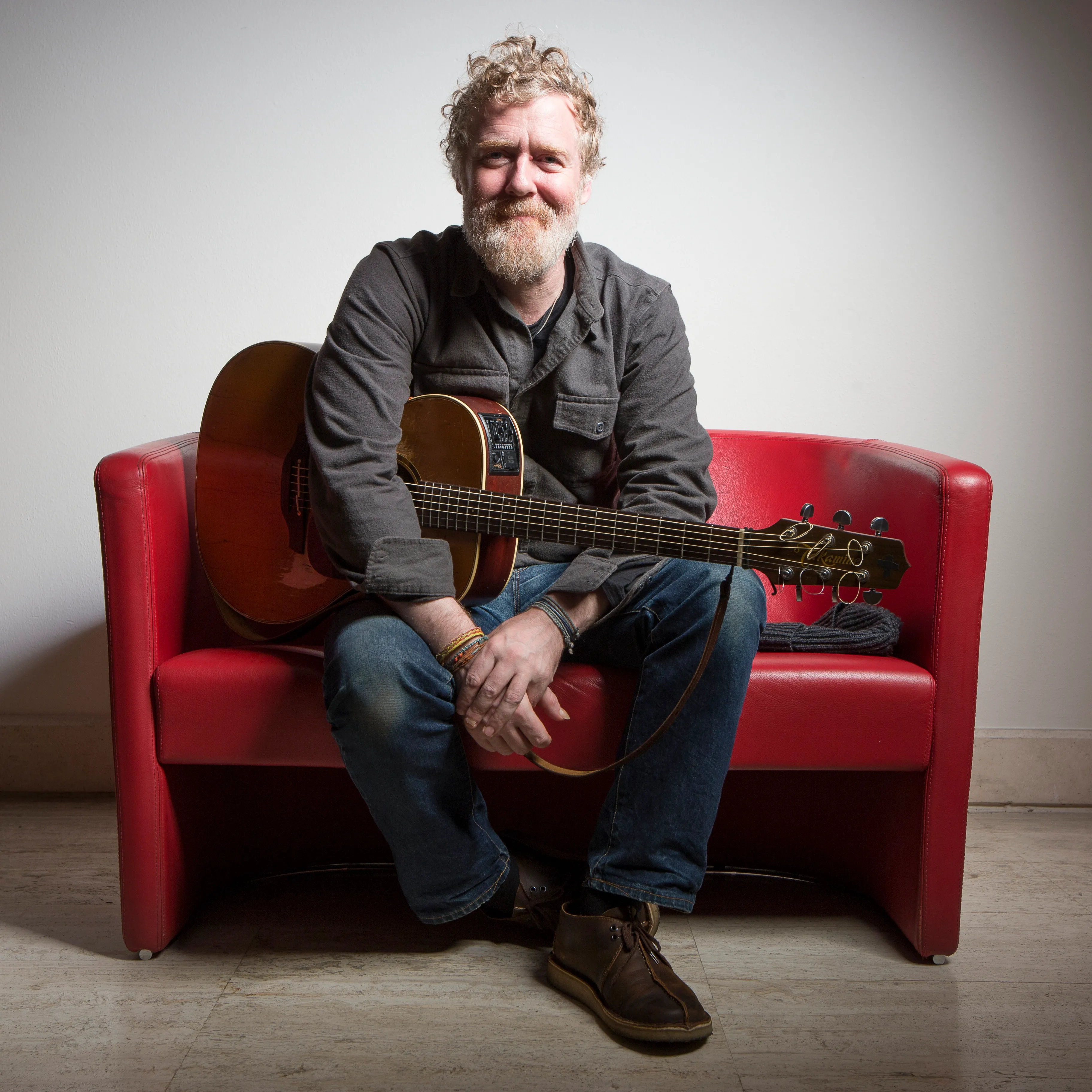 Irish musician Glen Hansard sitting on a red couch with an acoustic guitar.
