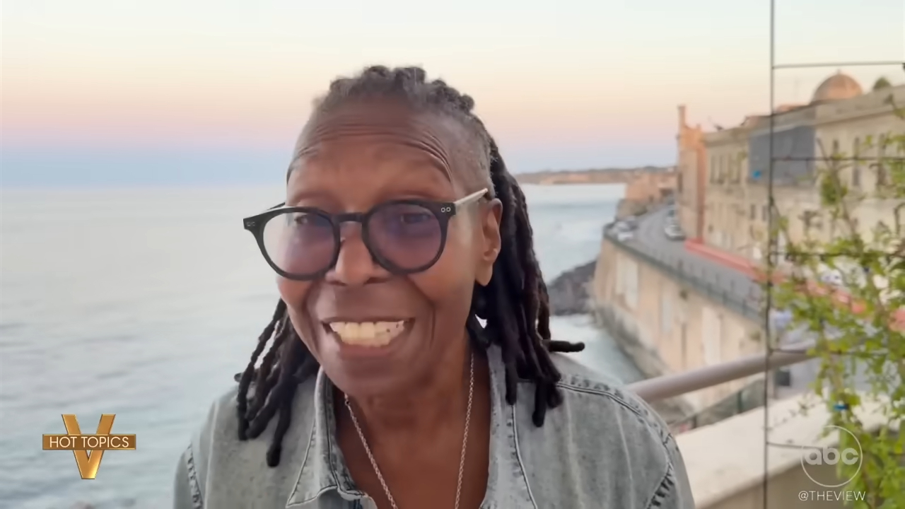 Whoopi Goldberg smiling in front of an ocean view with buildings along the coast.
