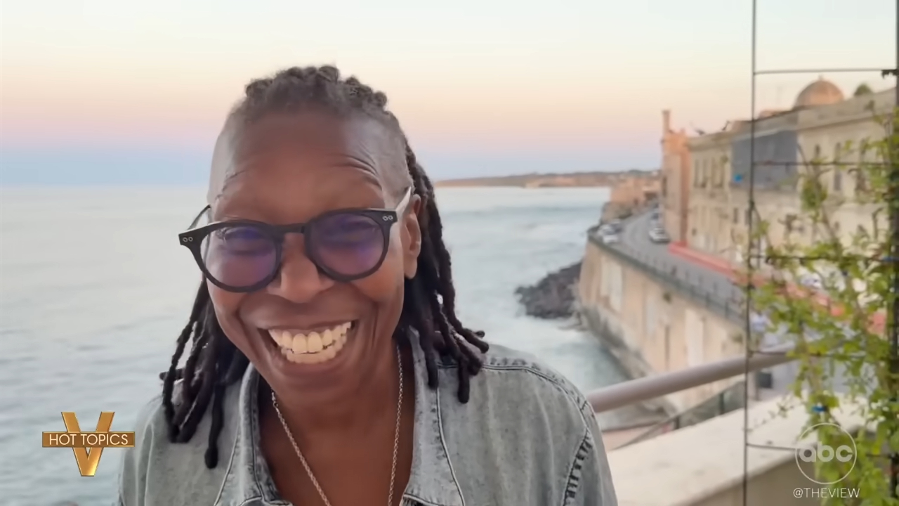 Whoopi Goldberg smiling and laughing with a coastal town in the background.