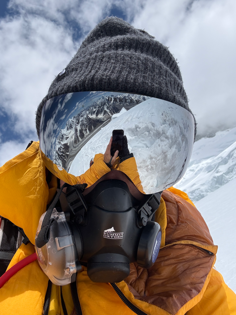 Napier Fuller wearing a beanie, reflective goggles, and an oxygen mask, on a snowy mountain.