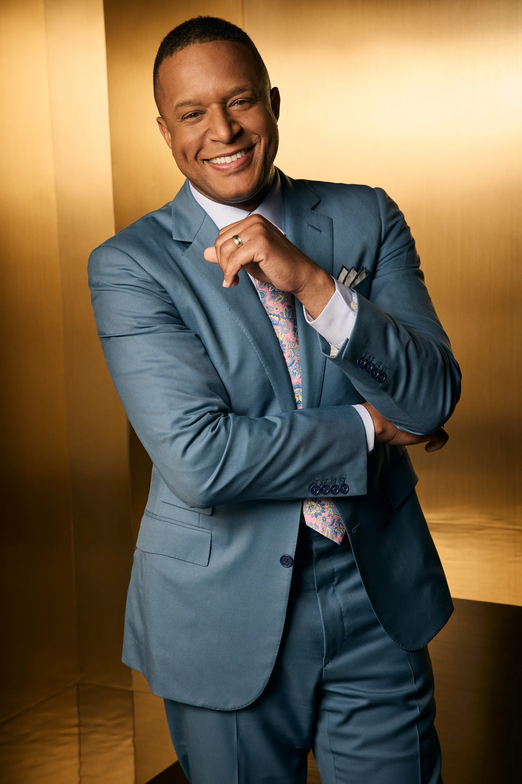 Craig Melvin posing in a teal suit with a patterned pink tie.