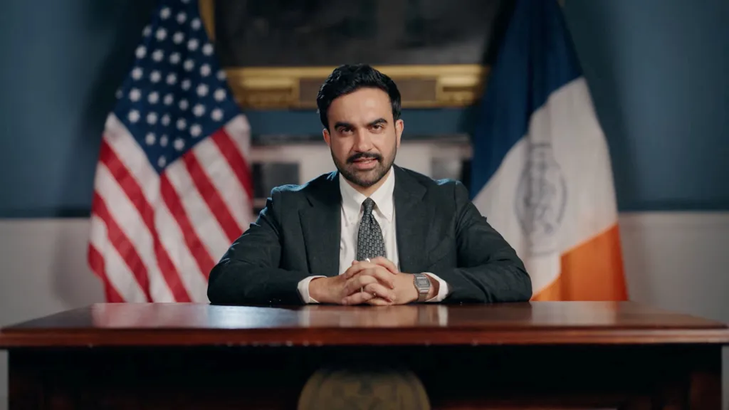 New York City Mayor Zohran Mamdani seated at a desk with an American flag and a New York City flag behind him.