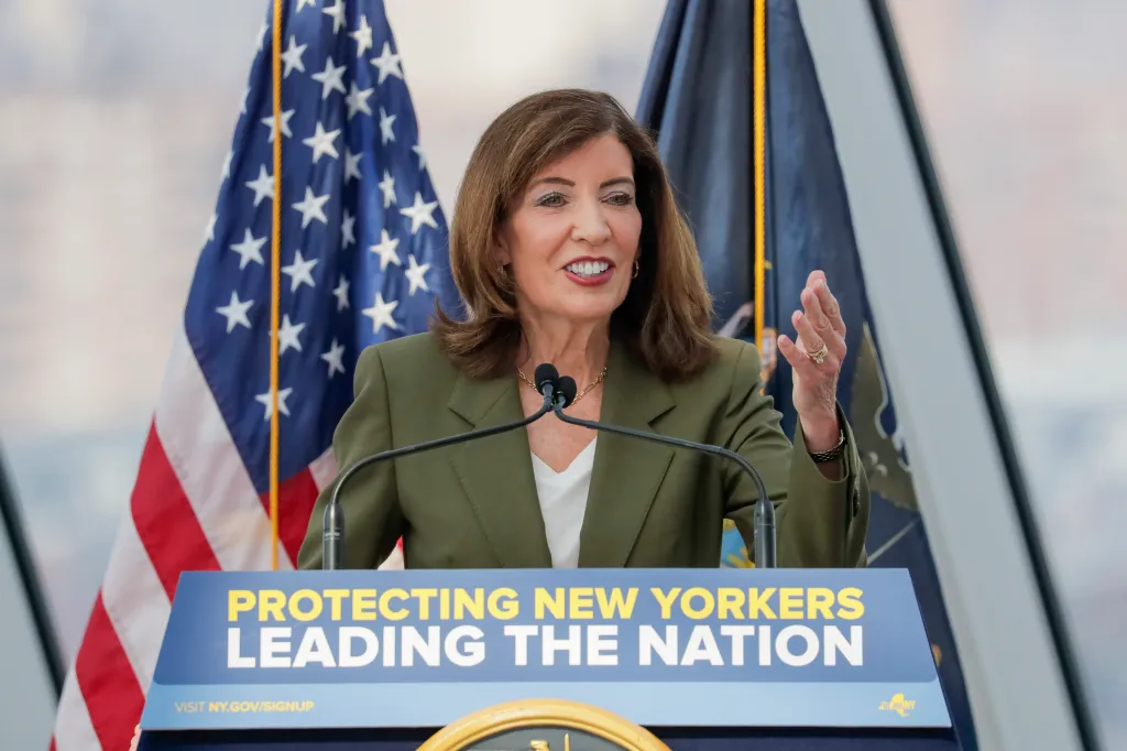 New York State Governor Kathy Hochul speaking at a podium with two flags behind her.