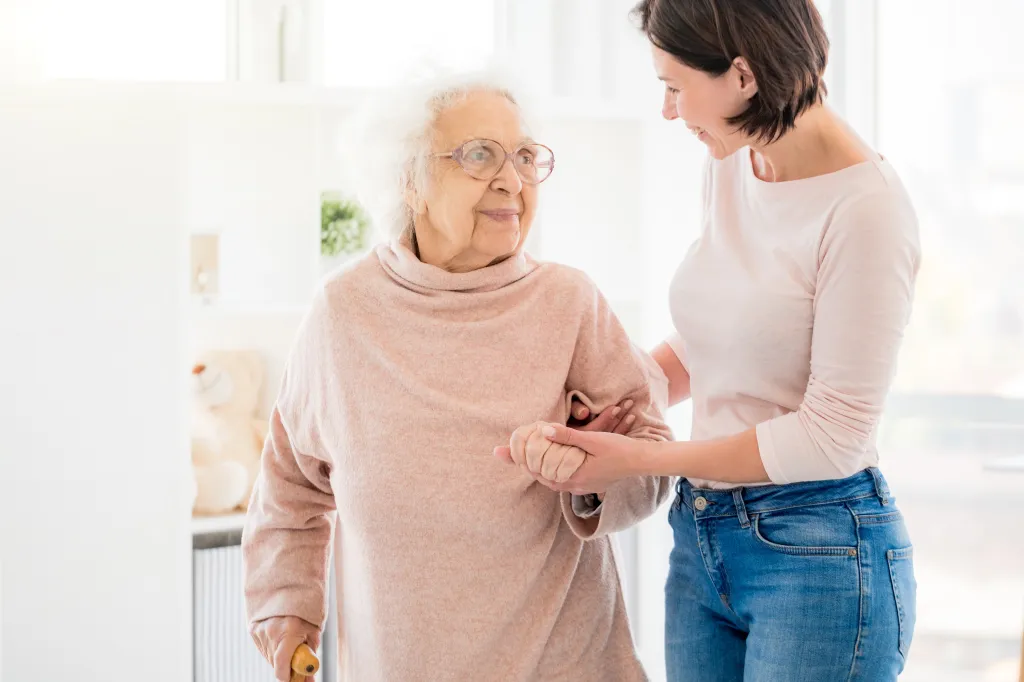 Young woman helping an elderly woman with white hair and glasses to walk.