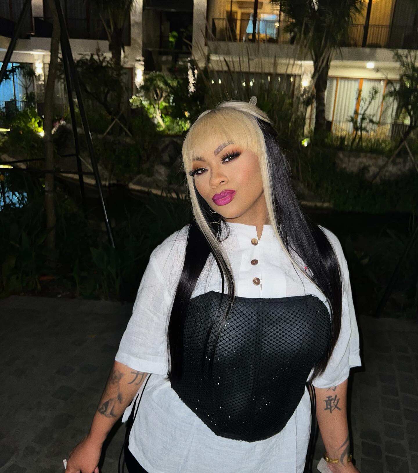 Nivea in a white shirt and black corset with long blonde and black hair and pink lipstick.