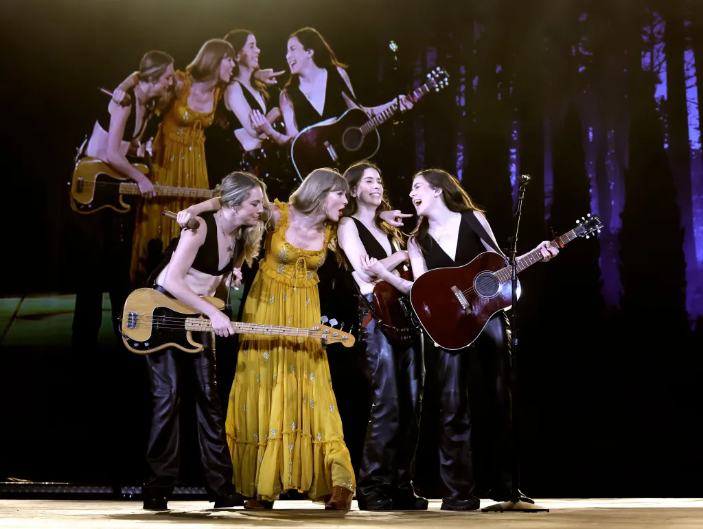 Taylor Swift and the band HAIM performing onstage with instruments and a video of themselves in the background.