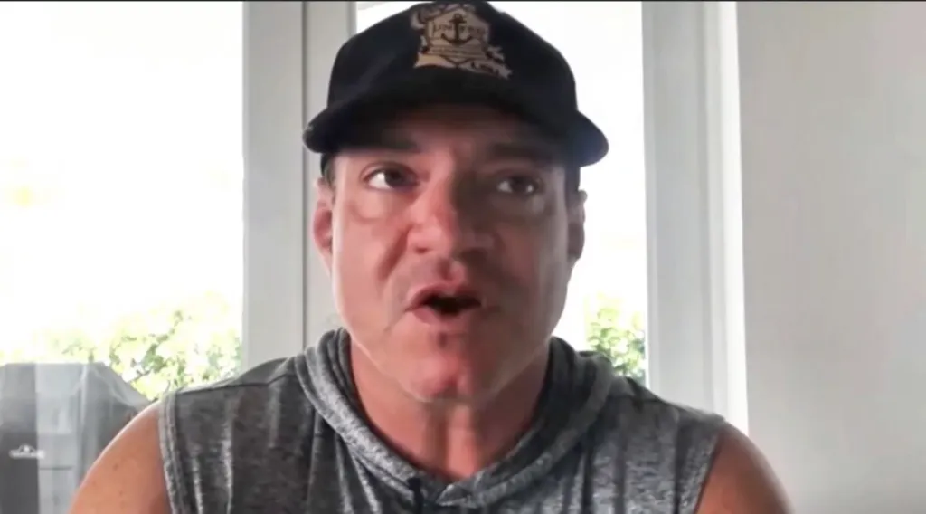 A man speaking to the camera, wearing a black cap with an emblem and a gray sleeveless hoodie.
