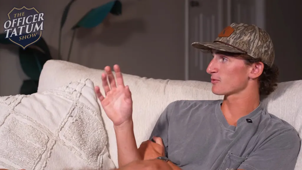 A man wearing a camouflage cap and grey t-shirt sits on a couch, gesturing with his hand during an interview for "The Officer Tatum Show".