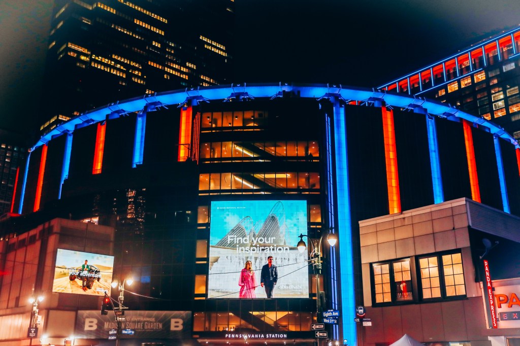 Madison Square Garden at night, lit with red and blue lights, with an advertisement for "Abu Dhabi" in the center.