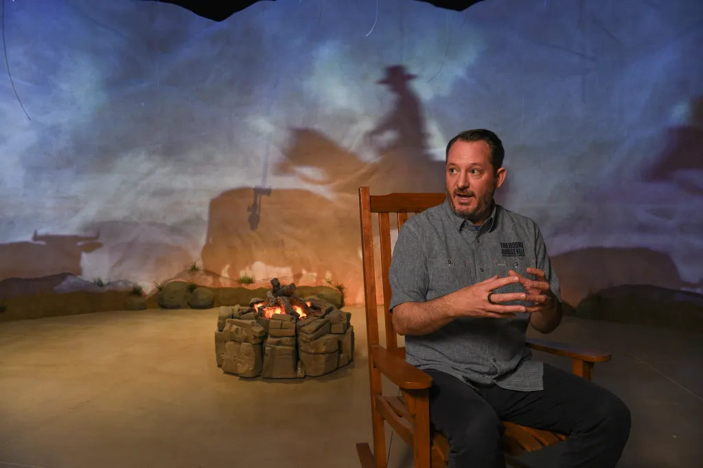 A man sits in a rocking chair in front of a campfire with a backdrop showing the shadow of a cowboy on a horse and a bison.