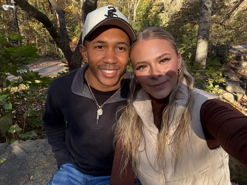 A man in a baseball cap and a woman with long blonde hair are posing for a selfie outdoors.