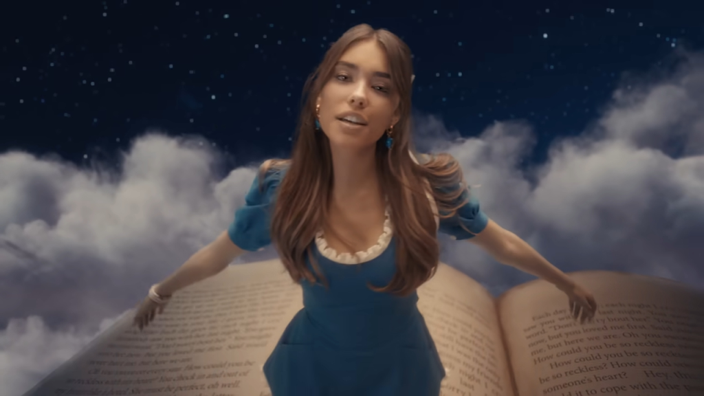 Madison Beer in a blue dress floating on a giant open book with clouds and a starry night sky in the background.