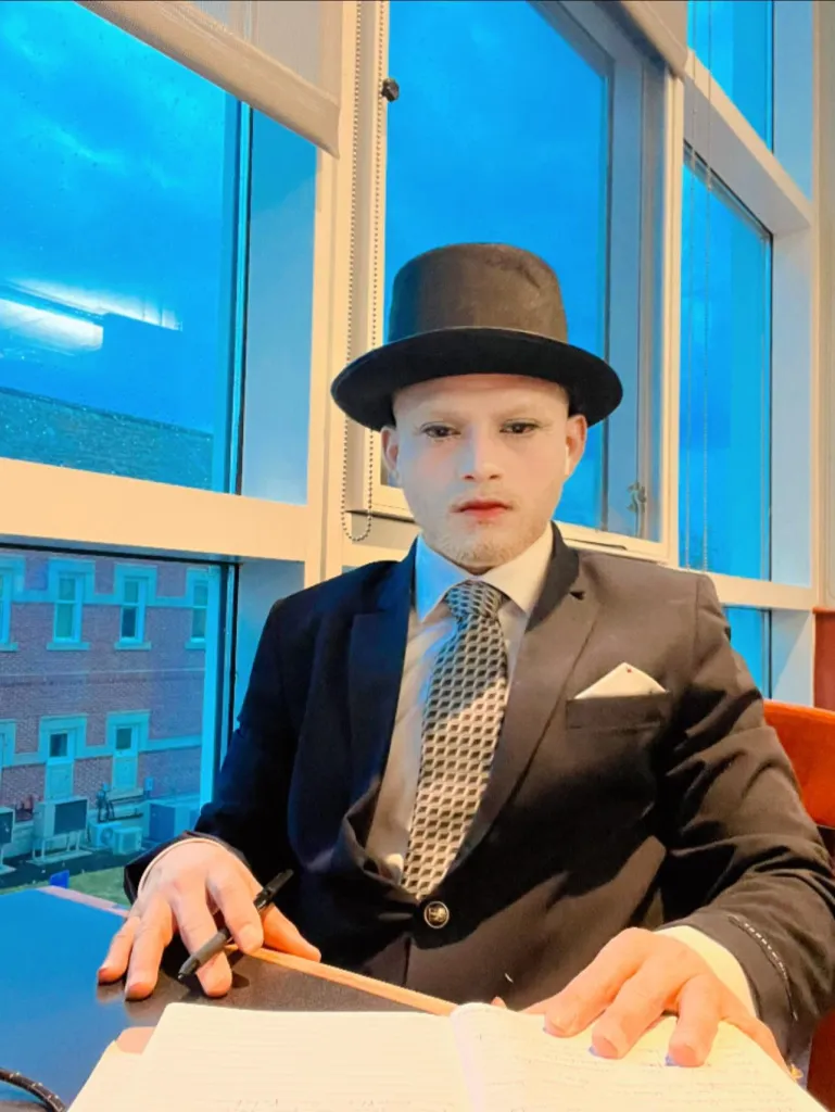 Hernan Villagran Lux in a suit and top hat with stage makeup, holding a pen over a notebook.