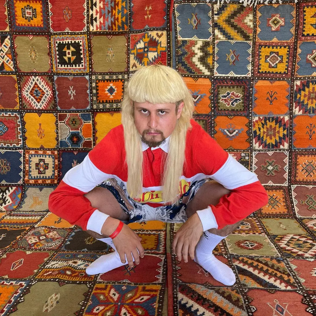 Oliver Tree crouching in front of a colorful, patterned rug.