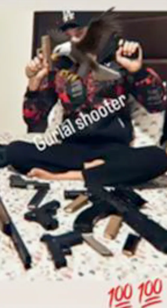 Person wearing a baseball cap holding two handguns, surrounded by an array of guns, magazines, and other weapons on a bed.