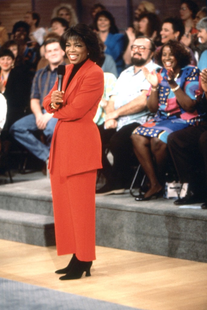 Oprah Winfrey in a red pantsuit on the set of her talk show, holding a microphone while a studio audience claps behind her.