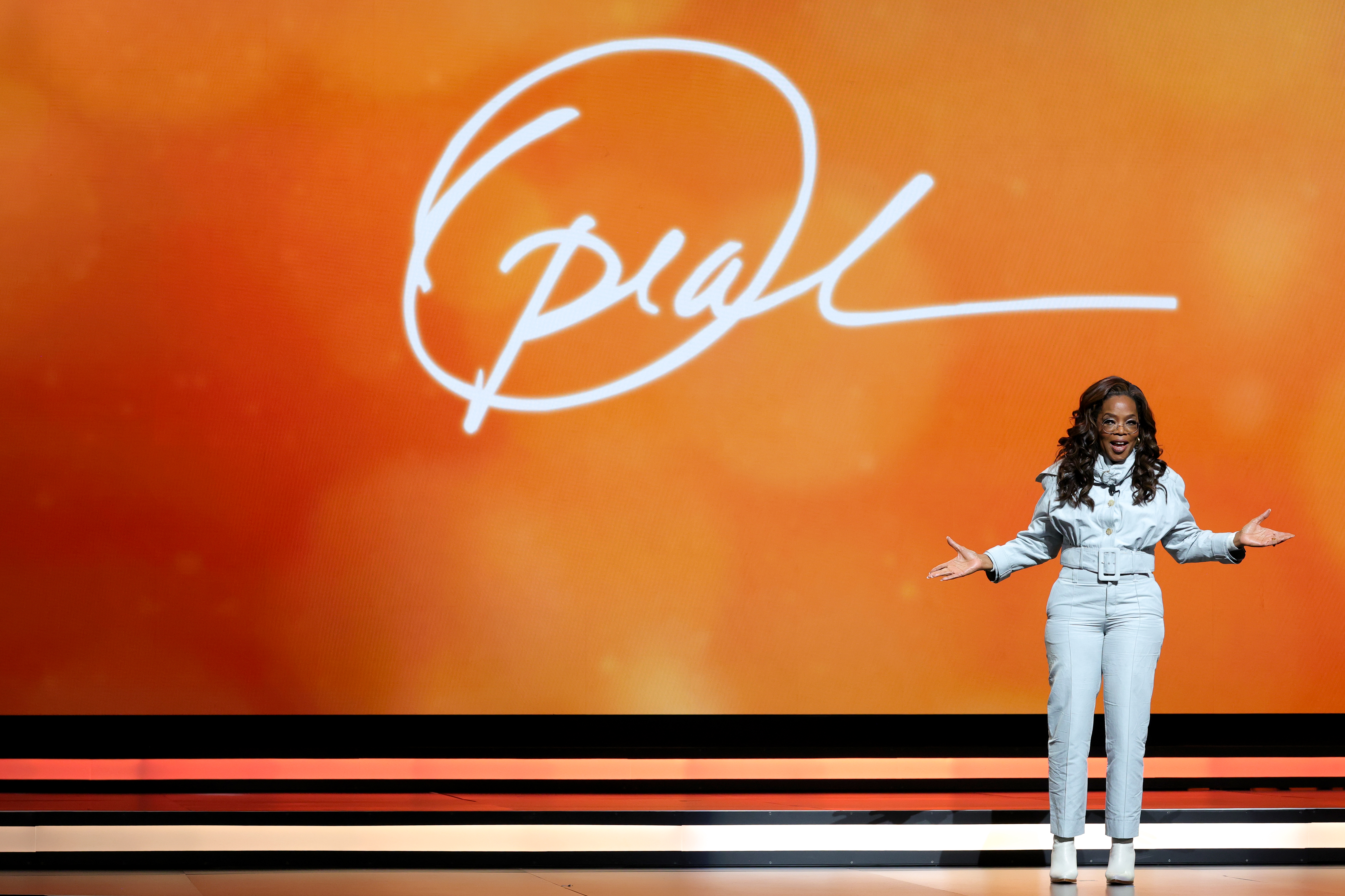 Oprah Winfrey speaks onstage with her name in white script on an orange background behind her.
