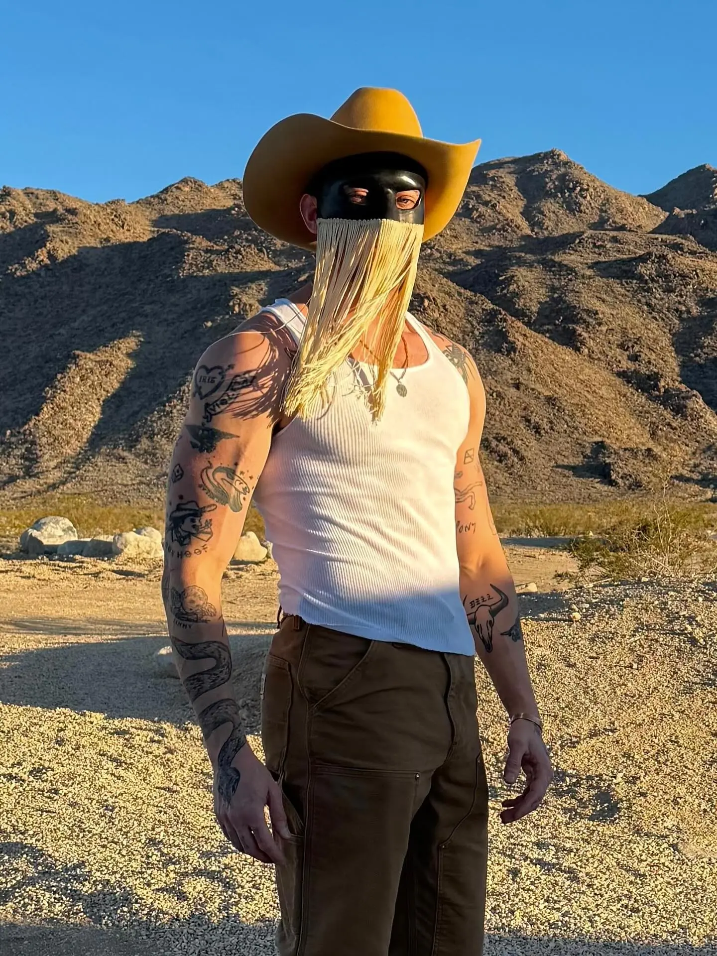 Orville Peck wearing a yellow cowboy hat and a black fringed mask, a white tank top, and brown pants in front of desert mountains.