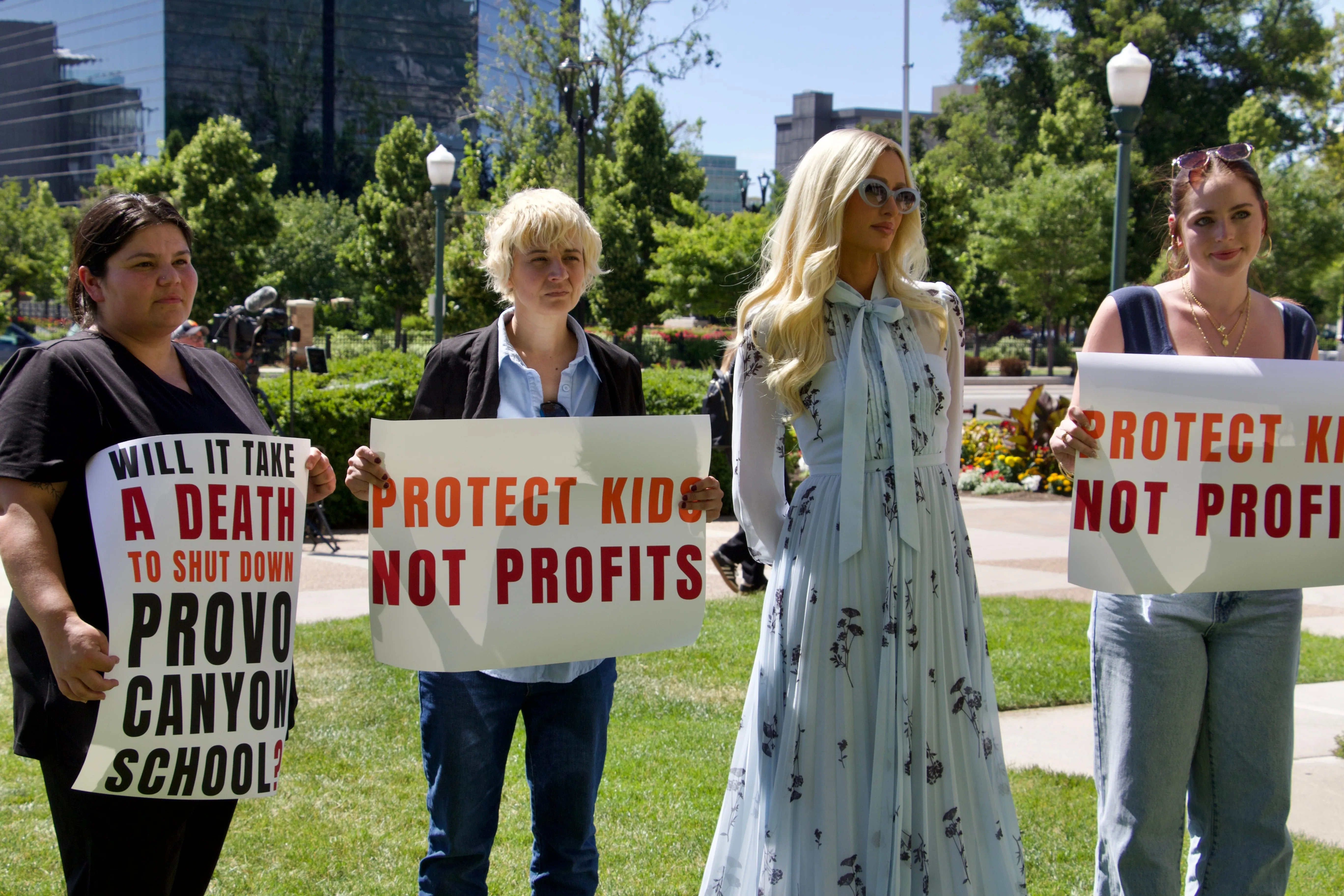 Paris Hilton with fellow survivors of residential teen treatment facilities holding signs that read "PROTECT KIDS NOT PROFITS" and "WILL IT TAKE A DEATH TO SHUT DOWN PROVO CANYON SCHOOL?"