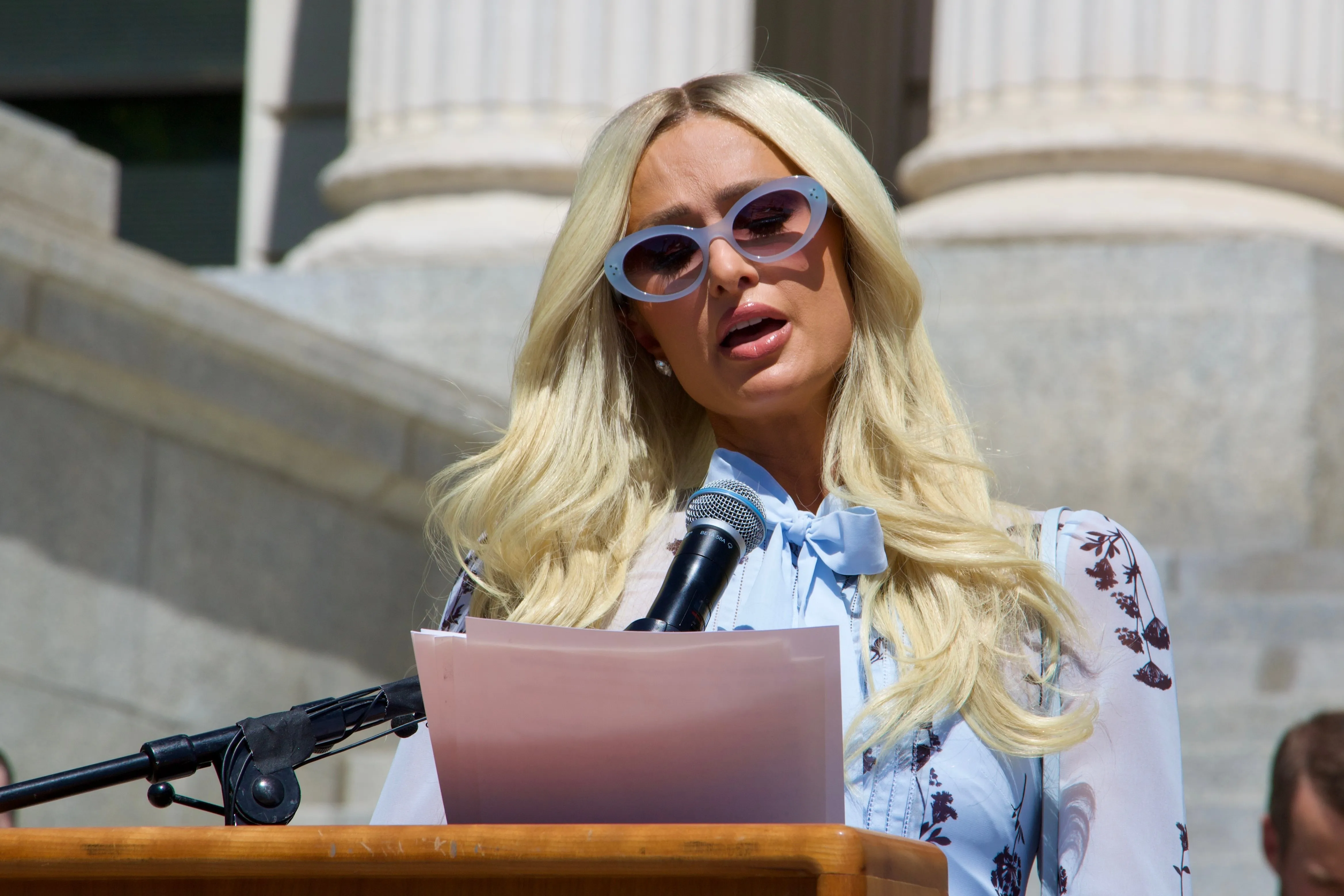 Paris Hilton speaks at a podium while wearing sunglasses and a light blue blouse.