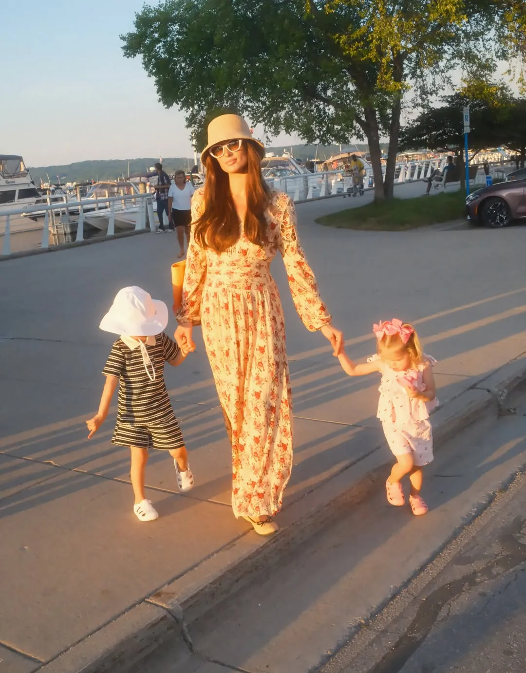 Paris Hilton with brunette hair, holding hands with two children by a marina.
