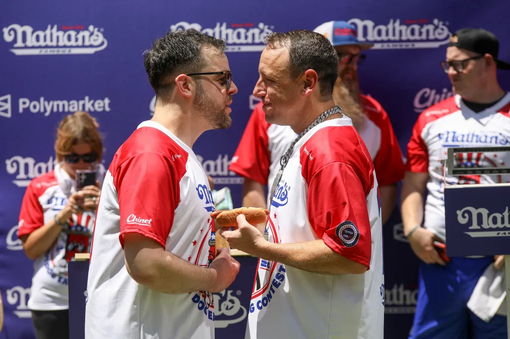 Joey Chestnut and Patrick Bertoletti face each other during a hot dog eating contest weigh-in, with Chestnut holding a hot dog.