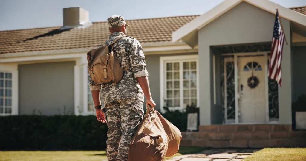 A soldier in camouflage uniform with a backpack and duffel bag walks toward a house with an American flag.