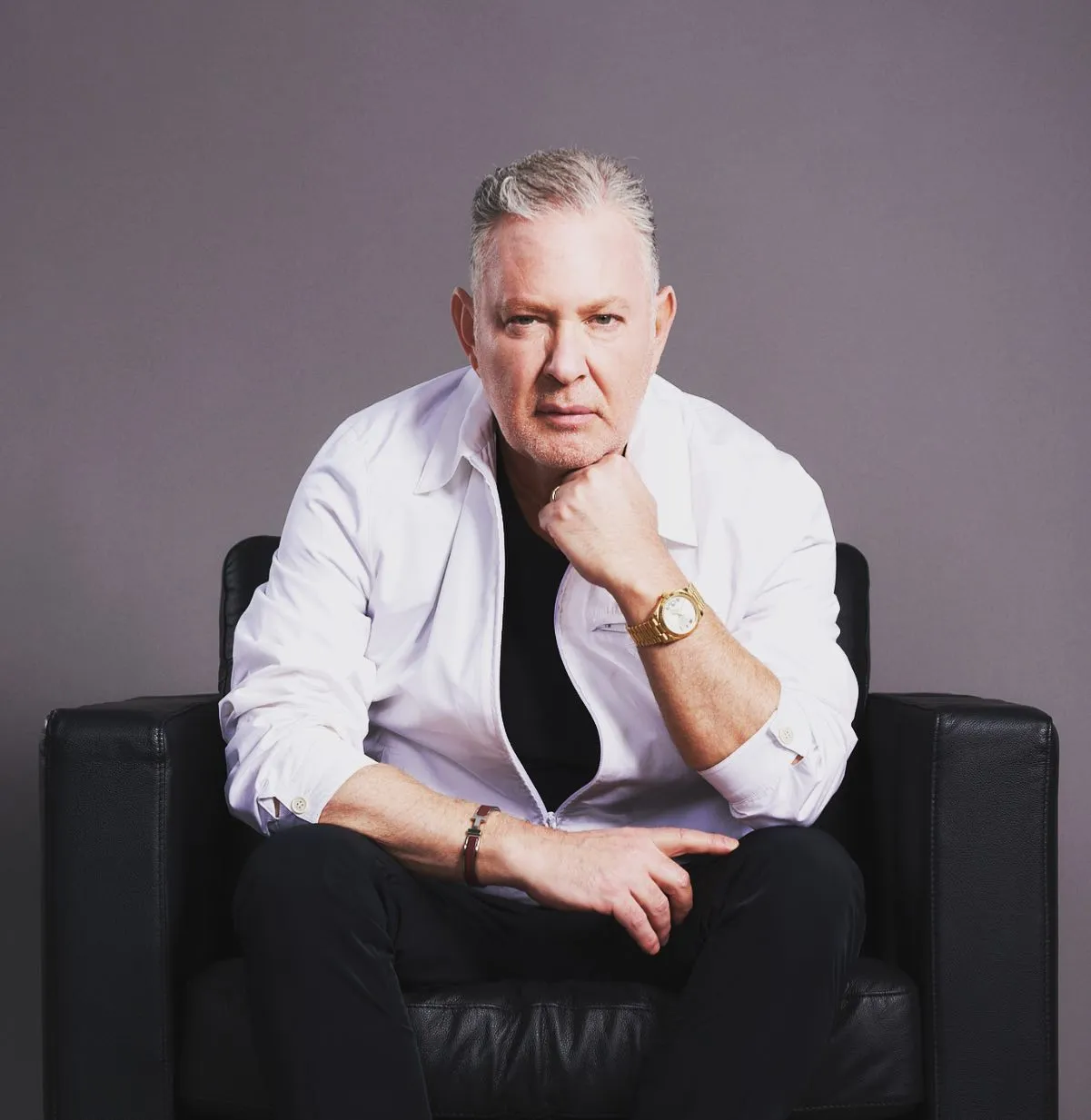 Paul Kemsley sitting in a black leather chair, wearing a white shirt and black t-shirt, with a gold watch and bracelet.