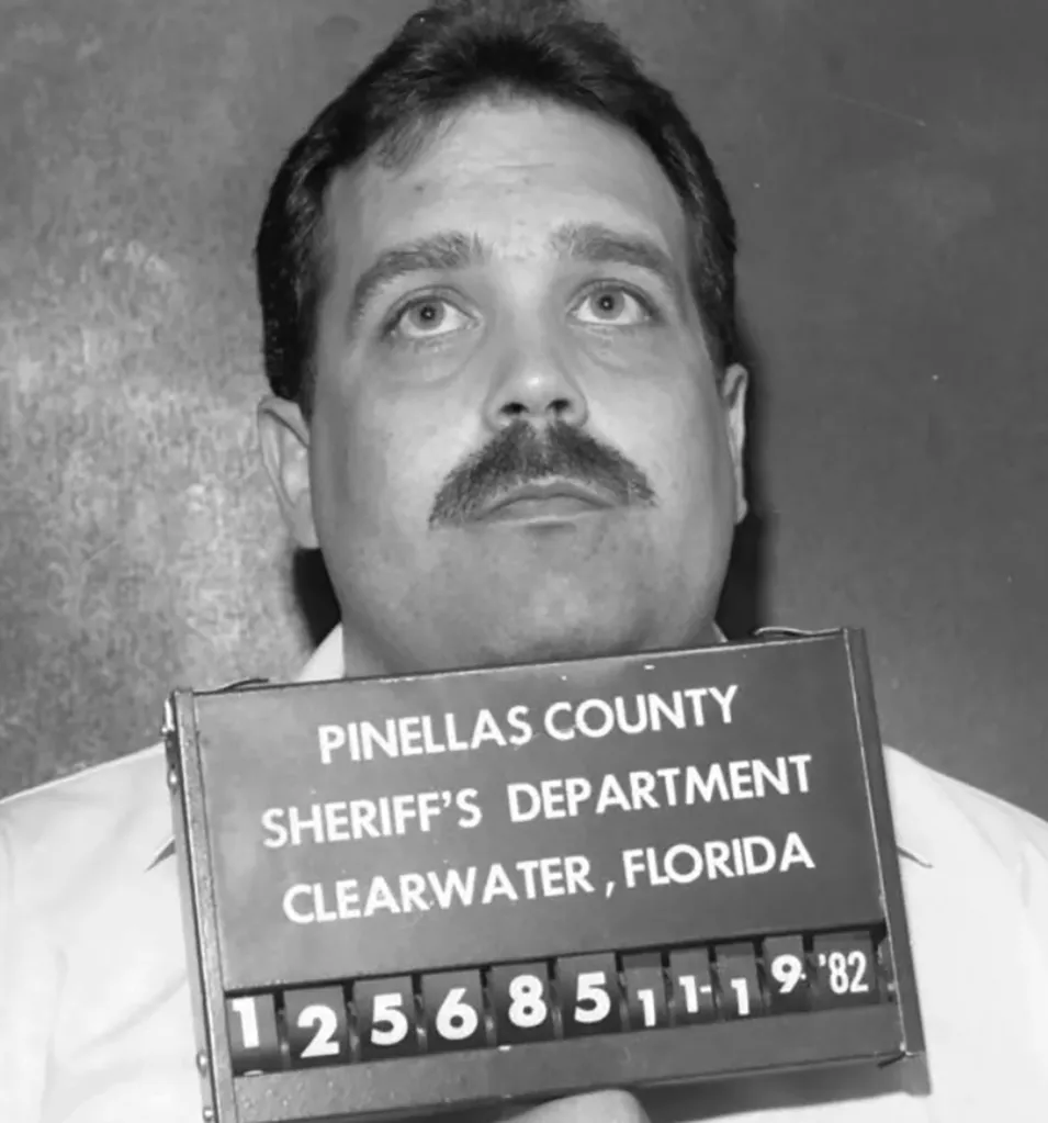 Booking photo of Paul Skalnik, a man with a mustache, looking up as he holds a mugshot board from the Pinellas County Sheriff's Department with the date 11-19-82.