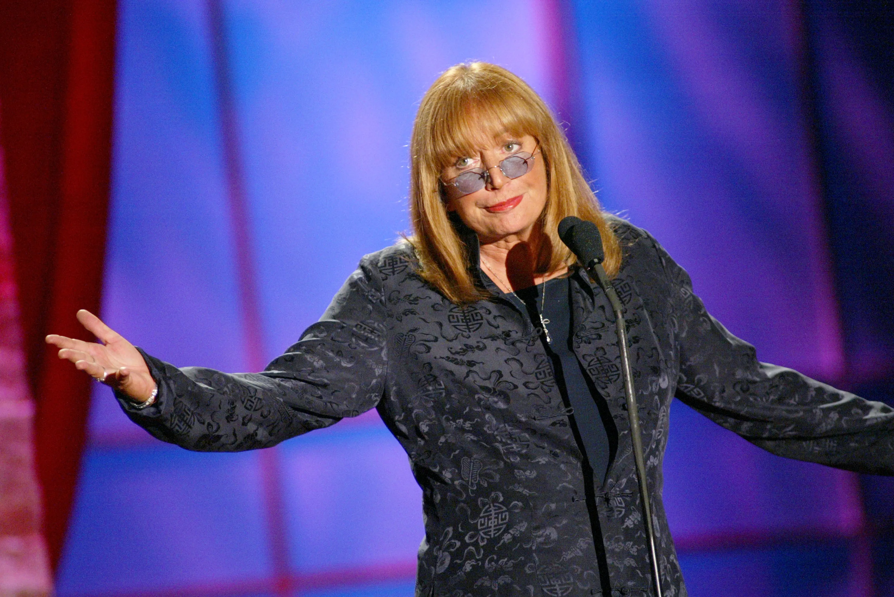 Penny Marshall presenting an award, standing at a microphone with arms outstretched, wearing a patterned black jacket and glasses.