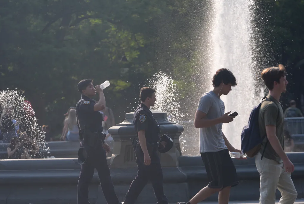Two police officers and two men walk past a fountain, with one officer drinking from a water bottle.