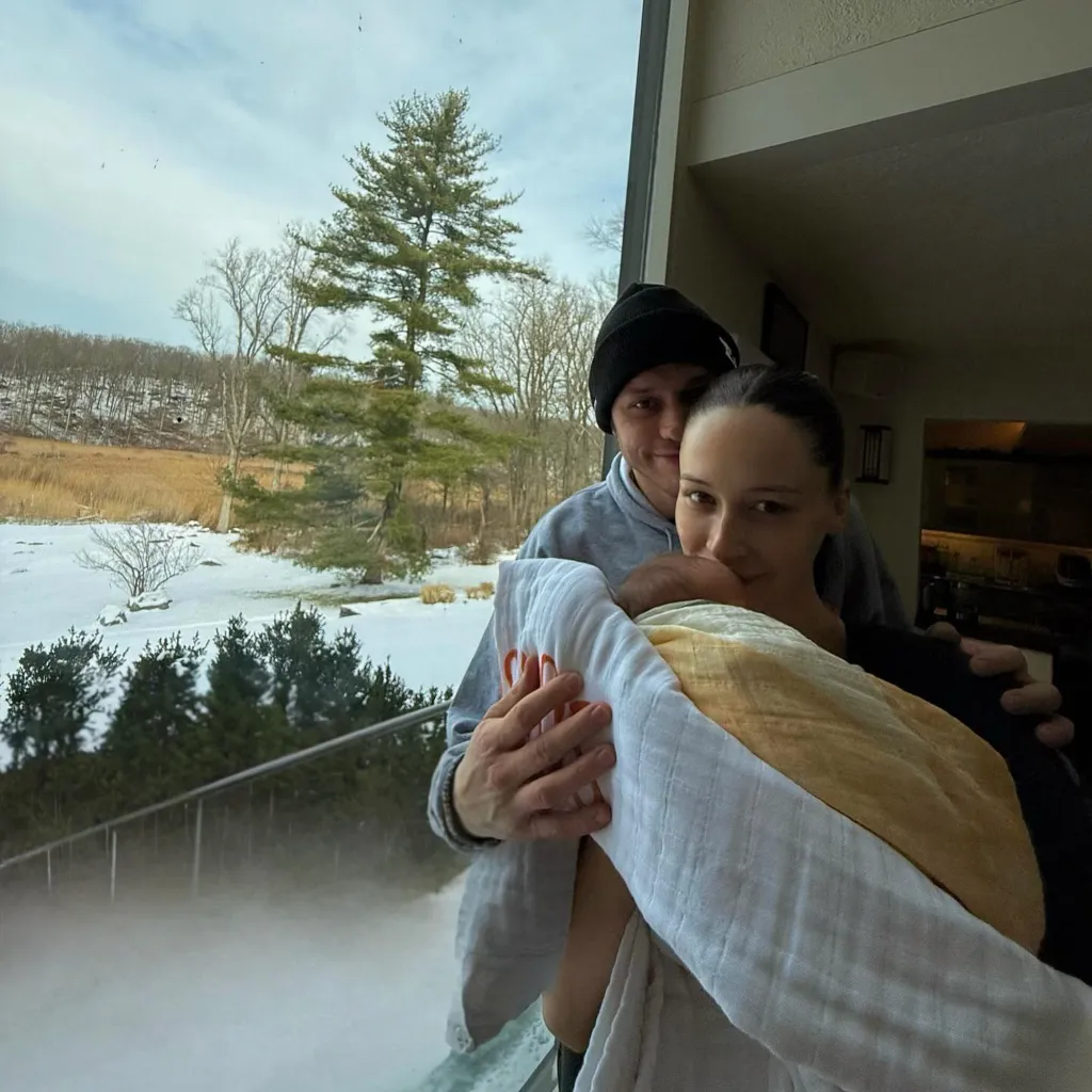 Pete Davidson and Elsie Hewitt hold their baby, swaddled in white and yellow blankets, in front of a snowy landscape.