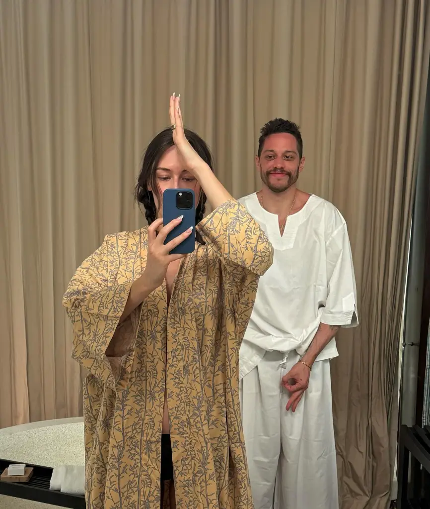 A woman holding a phone to her face taking a mirror selfie and a man standing behind her in white clothing.