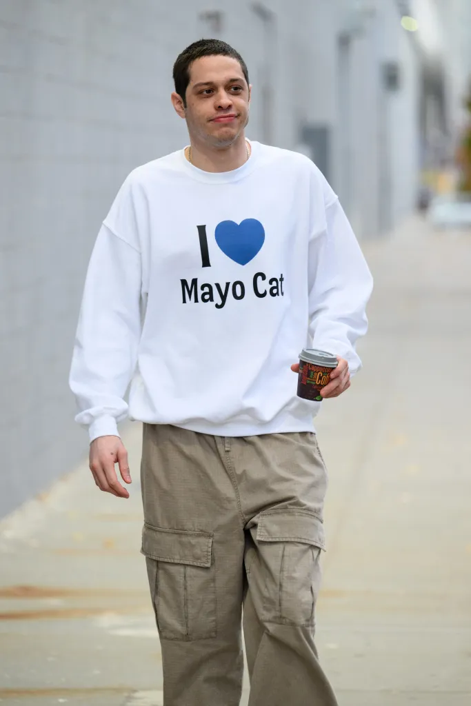 Pete Davidson walking while wearing a white "I (blue heart) Mayo Cat" sweatshirt and holding a coffee cup.