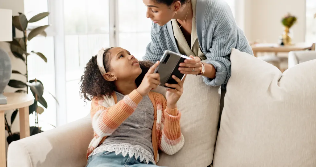 A mother taking a phone from her child on a couch to set screen time limits.