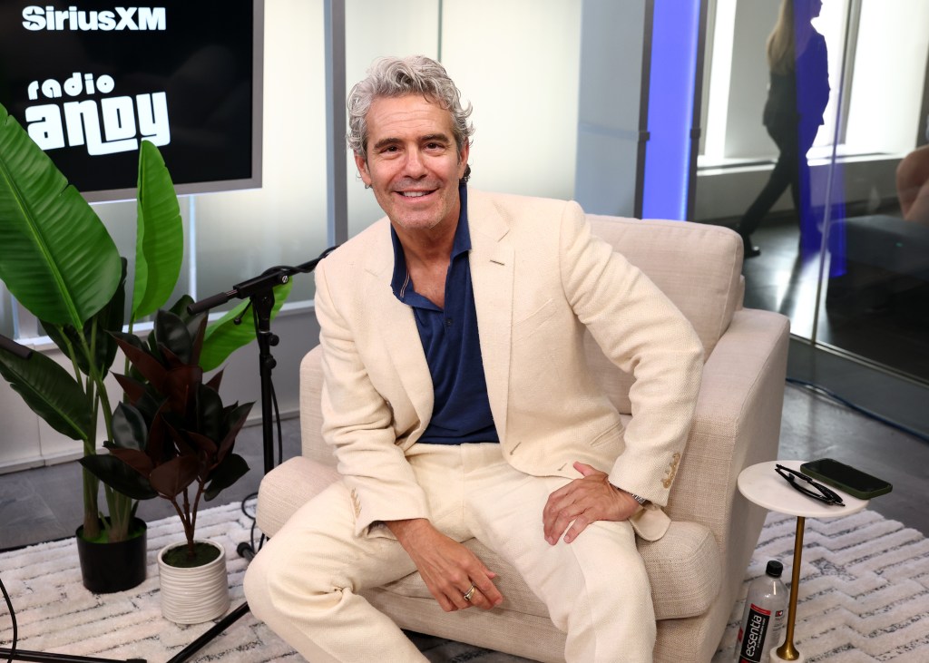 Andy Cohen smiling in a cream-colored suit on the set of SiriusXM's 