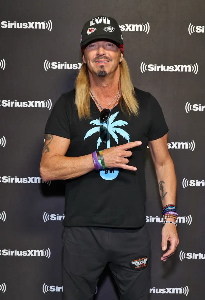 Bret Michaels poses in a black T-shirt and black Super Bowl hat, giving a rock-on gesture.