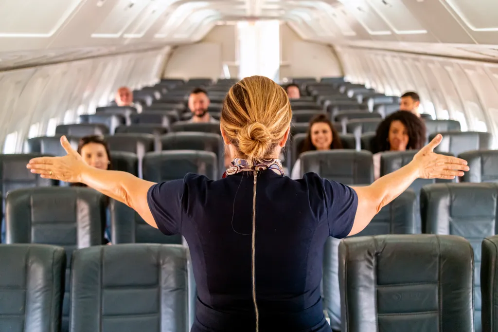 A flight attendant faces away from the camera, arms outstretched, demonstrating safety instructions to passengers in an airplane cabin.