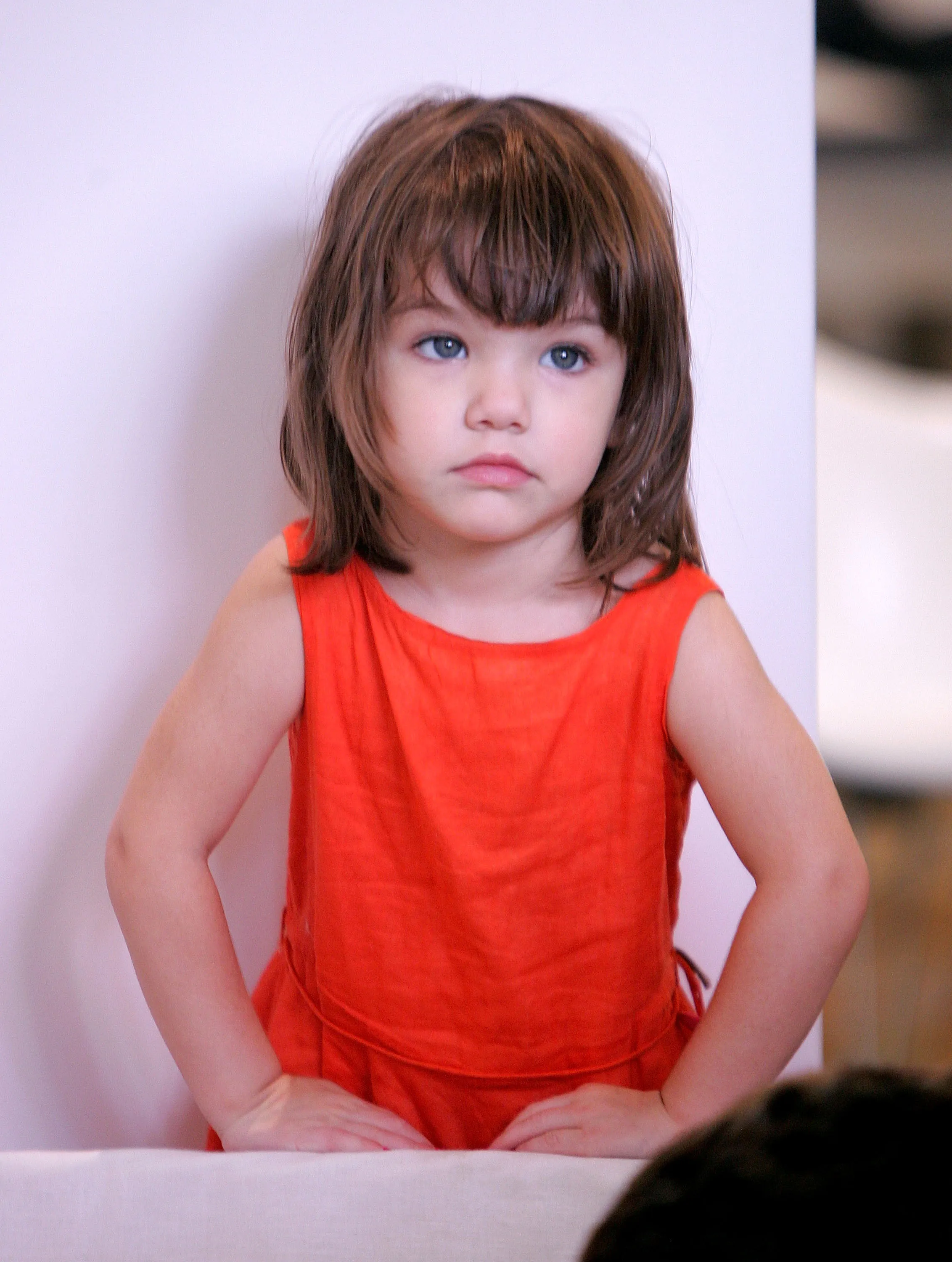Suri Cruise, a child with shoulder-length brown hair and a short fringe, looking up with wide, blue eyes, stands with her hands placed on a white surface, wearing an orange sleeveless top.