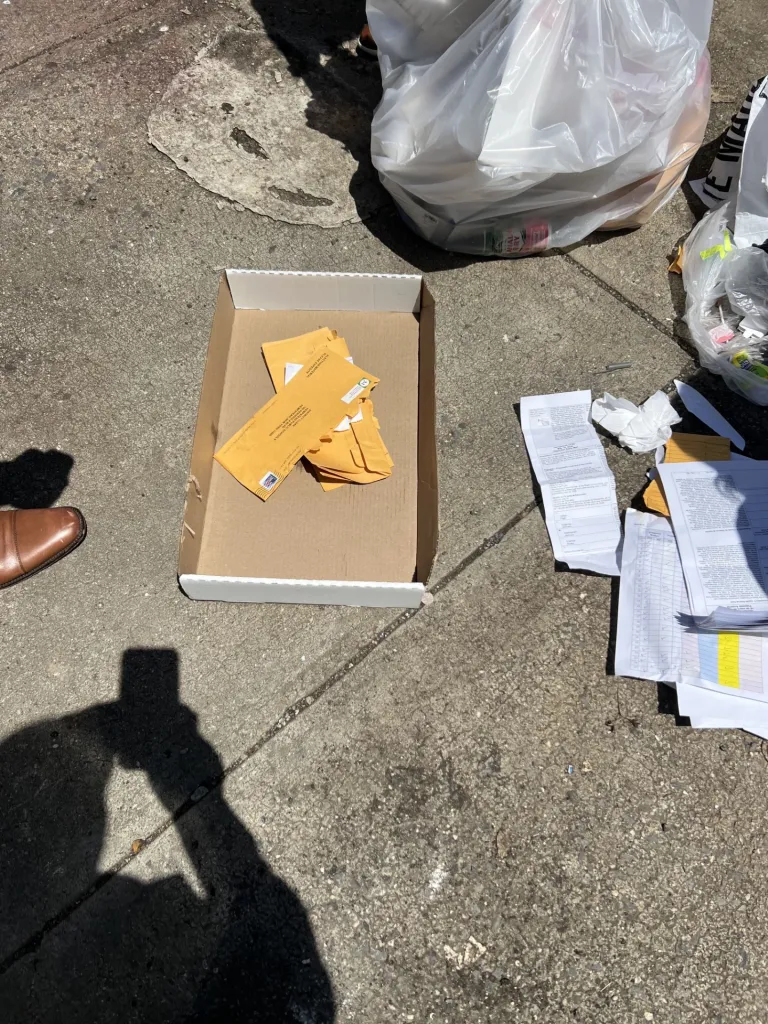 Election ballots and documents in a box on the ground next to a trash bag.