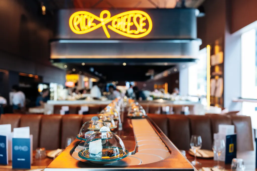 Pick & Cheese restaurant in Manhattan, with a cheese conveyor belt in the foreground and a neon sign in the background.